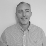 Black-and-white head-and-shoulders portrait of a person wearing a light-coloured button-down shirt, facing the camera against a plain light background. The image is cropped from the chest upwards and has a professional headshot style.